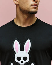 A man wears the Psycho Bunny MENS ARCHIVE BUNNY GRAPHIC TEE (MT0200031), a regular fit black shirt with a large graphic bunny—pink ears and skull face—on a pink background. Only his mouth, beard, and neck are visible in the image.