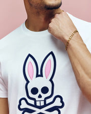 A person wearing the Psycho Bunny MENS ARCHIVE BUNNY GRAPHIC TEE (MT0200031), featuring a white Pima cotton shirt with a large skull and crossbones bunny ears graphic, gold chain bracelet, hand on chin, posed against a pink background.