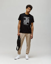 A man stands against a plain background wearing the Psycho Bunny MENS GLENN GRAPHIC TEE - MT0200034 with a bunny graphic, beige joggers, and white high-top sneakers. He holds a black bag in his left hand and looks relaxed.