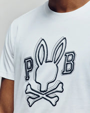 A man wears the Psycho Bunny MENS BALES GRAPHIC TEE (MT0200035), a Pima cotton shirt with a bold outlined Bunny graphic above crossed bones and "P" and "B" letters—ideal for year-round style.