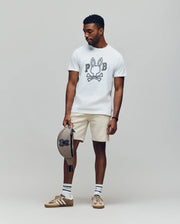 A man stands against a plain backdrop wearing Psycho Bunny’s MENS BALES GRAPHIC TEE (MT0200035) in white with a Bunny print, beige shorts, striped socks, brown sneakers, and holding a matching beige cap—ideal for year-round style.