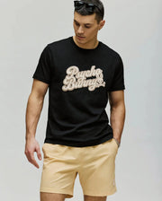 A man stands against a plain background wearing sunglasses, the Psycho Bunny MENS ELIAS GRAPHIC TEE (MT0200047) with a retro Bunny graphic and beige lettering, paired with light beige shorts, looking down with his left hand in his pocket.