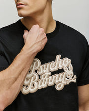 A man wears the Psycho Bunny MENS ELIAS GRAPHIC TEE (MT0200047), a black Pima cotton shirt with bold textured beige "Psycho Bunny" lettering and a retro bunny skull logo, partially covered by his raised left fist near his neck.