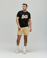 A man stands against a plain background wearing the Psycho Bunny MENS ELIAS GRAPHIC TEE (MT0200047) in black, paired with beige shorts, white sneakers and socks, and sunglasses on his head. He faces slightly to the side with one hand by his hip.