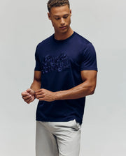 A man in light pants wears the MENS ELIAS GRAPHIC TEE (MT0200047) by Psycho Bunny—navy with embroidered script and a subtle Bunny graphic—standing against a plain light background, looking down with hands loosely clasped.