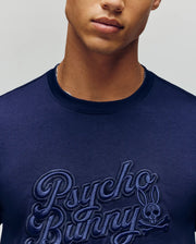 A man wears the MENS ELIAS GRAPHIC TEE (MT0200047) by Psycho Bunny—a navy Pima cotton shirt with an embroidered bunny skull logo and retro Bunny graphic, showing only his lower face and upper torso.