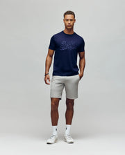 A man with short curly hair wears the MENS ELIAS GRAPHIC TEE - MT0200047 by Psycho Bunny, paired with light gray shorts, white socks, and white sneakers. He stands against a plain light background, relaxed with one hand in his pocket.
