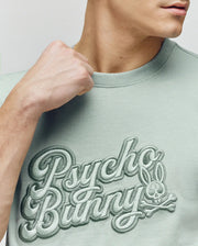 A person wears the Psycho Bunny MENS ELIAS GRAPHIC TEE - MT0200047, a light green Pima cotton shirt featuring textured "Psycho Bunny" embroidery and a retro Bunny graphic logo, with their fist raised and only their lower face showing.