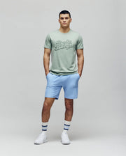 A man stands against a plain background wearing the Psycho Bunny MENS ELIAS GRAPHIC TEE - MT0200047 in light green, paired with light blue shorts, white socks with blue stripes, and white sneakers, his hands casually in his pockets.