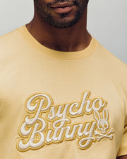 A man wears the Psycho Bunny MENS ELIAS GRAPHIC TEE (MT0200047) in pale yellow, featuring embroidered "Psycho Bunny" text and a bunny skull-and-crossbones graphic on the chest; only his lower face and upper torso are visible.