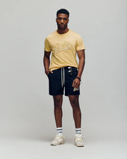 A man stands against a plain background wearing the Psycho Bunny MENS ELIAS GRAPHIC TEE - MT0200047 in yellow with a white Bunny graphic, black drawstring shorts, striped retro crew socks, white sneakers, and sunglasses hanging from his shorts pocket.