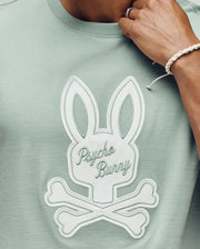 A person wears the Psycho Bunny MENS DEREK CENTER GRAPHIC TEE (MT0200048), a light green Pima cotton T-shirt with a large white embroidered bunny skull and crossbones logo and "Psycho Bunny" on the chest. Their ring-adorned hand is near their face.