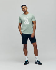 A man stands against a plain background in Psycho Bunny's MENS DEREK CENTER GRAPHIC TEE (MT0200048), a light green Pima cotton t-shirt with an embroidered white Bunny, paired with black shorts, white socks, black sneakers, and a relaxed pose.