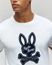 A person wearing the Psycho Bunny MENS DEREK CENTER GRAPHIC TEE (MT0200048), a white Pima cotton shirt featuring a large black embroidered bunny head above crossed bones with "Psycho Bunny" in cursive on the bunny's face.