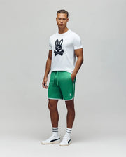 A man stands facing forward in a Psycho Bunny MENS DEREK CENTER GRAPHIC TEE - MT0200048, green athletic shorts with white trim, white socks with dark stripes, and white sneakers against a plain gray background.