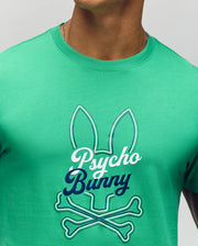 A person wears the Psycho Bunny MENS GARRET GRAPHIC TEE (MT0200050), a green crewneck made from soft Pima cotton, featuring a bunny ears outline with crossbones and bold "Psycho Bunny" script on the front.