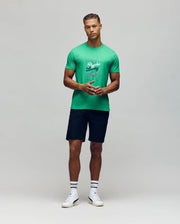 A man faces forward against a light gray background, wearing a bright green Psycho Bunny MENS GARRET GRAPHIC TEE (MT0200050), dark shorts, white crew socks, and white sneakers.