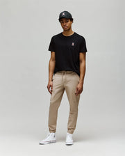 A man wears the black Psycho Bunny MENS MISCHIEF BUNNY TEE (MT0200051) in soft Pima cotton with an embroidered Mischief Bunny on the chest, paired with beige joggers and white high-top sneakers against a light gray background.