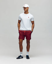 A man stands against a plain background wearing a white Psycho Bunny MENS MISCHIEF BUNNY TEE (MT0200051), red patterned drawstring shorts, striped white socks, and black sneakers. His hands are in his shorts' pockets.