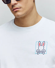 A person in Psycho Bunny’s MENS JESSE GRAPHIC TEE - MT0200056: a white Pima cotton shirt with a pink-eared bunny head over crossed bones, printed using eco-friendly water-based inks.