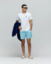 A man wearing black sunglasses, blue patterned shorts, beige slides, and holding a dark blue jacket stands against a plain gray background. He wears Psycho Bunny’s MENS JESSE GRAPHIC TEE (MT0200056), made of white Pima cotton with an eco-friendly print.