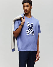 A young man stands in front of a plain light background, holding a jacket and wearing the Psycho Bunny MENS RYDER OVERSIZED FIT TEE (MT0200062), a loose Pima cotton shirt with a bold bunny skull and crossbones graphic, paired with dark pants.