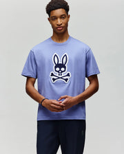 A young man wears the Psycho Bunny MENS RYDER OVERSIZED FIT TEE (MT0200062), a soft Pima cotton shirt with a large cartoon skull and crossbones topped with bunny ears, standing against a plain light background.