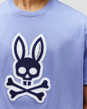 A person wears the Psycho Bunny MENS RYDER OVERSIZED FIT TEE (MT0200062), a light purple premium Pima cotton shirt featuring a bold graphic bunny with skull and crossbones, outlined ears, and facial details in white and black.