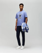 A man stands against a plain background wearing the Psycho Bunny MENS RYDER OVERSIZED FIT TEE (MT0200062), a light purple premium Pima cotton shirt with an embroidered Bunny skull and crossbones, dark pants, white sneakers, and holds a matching jacket.
