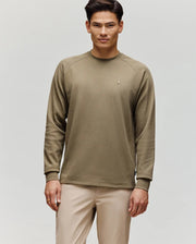 A man with short dark hair wears the Psycho Bunny MENS EVANS WAFFLE - MT0200091 light olive green cotton knit long-sleeve sweatshirt and beige pants, standing against a plain light grey background, facing forward neutrally.