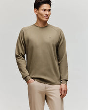A man with short, styled hair and a slight smile stands against a plain background, wearing Psycho Bunny’s MENS EVANS WAFFLE - MT0200091 khaki cotton knit crewneck sweater and light beige pants, one hand in his pocket.