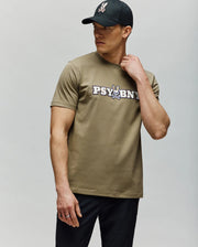 A man in a black cap and the Psycho Bunny MENS WADE GRAPHIC TEE (MT0200105)—olive-green with "PSYBUNY" and a cartoon bunny graphic—stands against a light gray background, looking sideways with one hand on his neck.