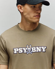 A man wears a Psycho Bunny MENS WADE GRAPHIC TEE (MT0200105) in beige, featuring "PSYBNY" and a bunny skull and crossbones on the chest, made from 100% cotton in a regular fit, paired with a black cap, looking to his left.