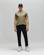 A man stands against a plain background in the Psycho Bunny MENS WADE GRAPHIC TEE - MT0200105, a khaki 100% cotton regular fit t-shirt, paired with a black cap, black pants, and light sneakers. He faces forward with a neutral expression.
