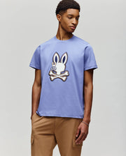 A young man wears the Psycho Bunny MENS WADE GRAPHIC TEE (MT0200106), a 100% cotton shirt with a large embroidered bunny head above crossed bones, paired with tan pants, standing against a plain light gray background.