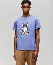 A young man with short hair models the Psycho Bunny MENS WADE GRAPHIC TEE (MT0200106), a 100% cotton shirt featuring an embroidered bunny above crossed bones, paired with tan pants against a light gray background.