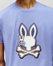 A person wearing the Psycho Bunny MENS WADE GRAPHIC TEE (MT0200106), a light purple 100% cotton shirt with a large white bunny skull and crossbones graphic, plus a smaller bunny skull and crossbones on the cheek.
