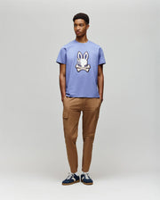 A young man wears the Psycho Bunny MENS WADE GRAPHIC TEE - MT0200106 in lavender, featuring a 100% cotton build and an embroidered bunny, paired with tan cargo pants and dark sneakers against a plain background.