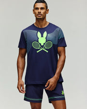 A man wears the Psycho Bunny TENNIS BALL CREW MENS TEE ENGLISH (MT0200113) in navy with neon green Tennis Bunny rackets and a stylized bunny logo, paired with matching navy shorts, standing against a light gray background.