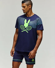 A man wears a Psycho Bunny TENNIS BALL CREW MENS TEE ENGLISH (MT0200113), a navy blue Pima cotton tee with neon green crossed rackets and bunny logo, paired with shorts, standing against a plain light background.
