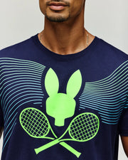A man wears the Psycho Bunny Tennis Ball Crew Men's Tee (MT0200113) in navy blue, made from soft Pima cotton, with a neon green bunny head above crossed tennis rackets and light blue radiating lines; only his lower face and upper torso show.