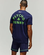 A man faces away in a navy Psycho Bunny TENNIS BALL CREW MENS TEE with “PSYCHO BUNNY” and a Tennis Bunny logo in neon green on the back, paired with shorts featuring green and blue stripes at the hem.