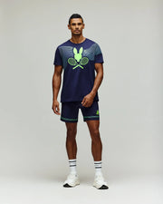 A man faces forward wearing the Psycho Bunny TENNIS BALL CREW MENS TEE ENGLISH (MT0200113), a navy Pima cotton tee with neon green tennis graphics, white athletic shoes, and white socks with navy stripes, against a plain light gray background.