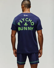 A man faces away in Psycho Bunny's TENNIS BALL CREW MENS TEE FRENCH (MT0200114), a navy Pima cotton tee featuring neon green "PSYCHO BUNNY" text and bunny logo, paired with coordinating navy shorts with green stripes.