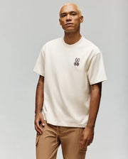 A man with short blond hair wears the Psycho Bunny MENS WADE OVERSIZED FIT CHEST TEE (MT0200152) in cream, made from soft Pima cotton with a small Bugs Bunny graphic on the left chest, paired with tan pants against a light gray background.