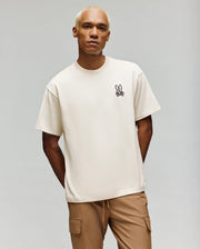 A person with short blond hair wears the Psycho Bunny MENS WADE OVERSIZED FIT CHEST TEE (MT0200152) in off-white, featuring a small cartoon bunny emblem, paired with tan cargo pants against a plain light background.