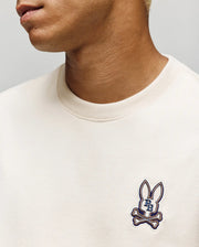 A person is seen wearing the Psycho Bunny MENS WADE OVERSIZED FIT CHEST TEE (MT0200152), a light beige, relaxed crewneck tee with a "BB" bunny head and crossbones patch on the left chest; only their lower face and neck show.