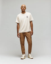 A person with light blond hair wears a Psycho Bunny MENS WADE OVERSIZED FIT CHEST TEE (MT0200152) in beige, styled with tan cargo pants and beige sneakers, standing against a plain light gray background.