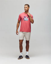 A man with a neutral expression wears the Psycho Bunny MENS SHANE GRAPHIC TEE - MT0200175, featuring a pink Pima cotton martini glass print, paired with light beige shorts, white socks, and black sneakers; one hand touches his neck.
