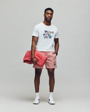 A man stands against a plain background wearing the Psycho Bunny MENS SHANE GRAPHIC TEE - MT0200175, red and white striped shorts, white sneakers, and a watch. He holds a folded pink towel under one arm and looks calmly at the camera.